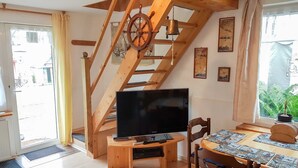A room with a wooden staircase, a television on a wooden stand, a dining table with placemats, and a window with a view of greenery. - Apartment 1 - family-friendly apartments in Recknitztal (Semlow)