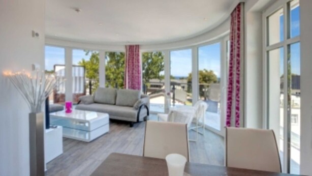 A modern living room with a sofa, coffee table, and chairs, large windows with curtains, and a view of greenery outside. - Villa Lara Whg.11, LAKE VIEW, ROOF TERRACE, CHAIR - Villa Lara, NEAR THE BEACH, (Heringsdorf)
