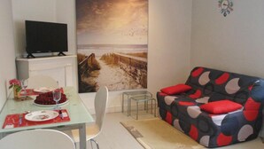 A living room with a large beach painting, a TV on a stand, a dining table with plates and glasses, and a colorful sofa. - Gorgeous apartment in Vildé-la-Marine (Vildé-la-Marine)