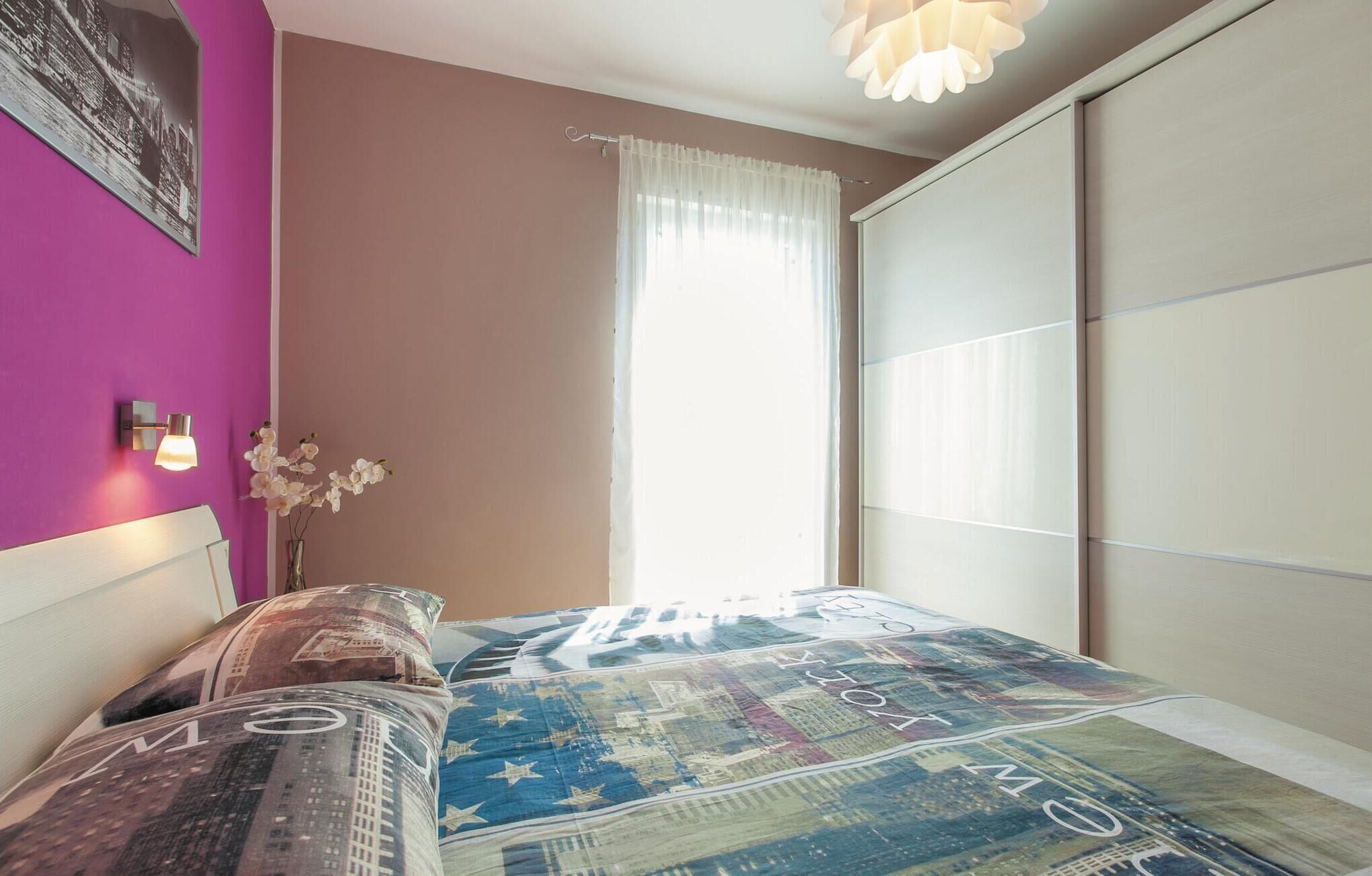 A bedroom with a bed, a nightstand, a vase with flowers, a window with sheer curtains, and a cityscape bedspread.