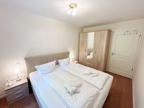 A neatly made bed with white linens and pillows, a bedside lamp, a wooden nightstand, and a large wardrobe with a mirror.
