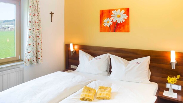 A hotel room with a bed, two pillows, a nightstand, a wall-mounted light, a painting, a window with curtains, and a radiator.