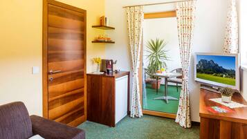 Une chambre d’hôtel avec une porte en bois, un petit bureau avec une télévision, un canapé, une petite table avec une plante, et une vue sur un espace extérieur verdoyant, visible à travers une porte vitrée.