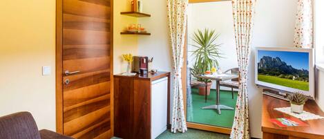 Habitación de hotel con puerta de madera, un escritorio pequeño con televisor, un sofá, una mesita con una planta y vista a un espacio verde al exterior a través de una puerta de vidrio.