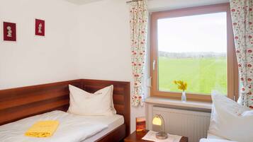 Une chambre à coucher avec un lit en bois, une table de chevet avec une lampe, une fenêtre ornée de rideaux à motifs floraux et une vue sur un champ verdoyant.