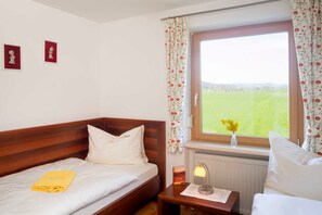 Une chambre à coucher avec un lit en bois, une table de chevet avec une lampe, une fenêtre ornée de rideaux à motifs floraux et une vue sur un champ verdoyant.