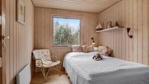 Une chambre confortable au décor intérieur en bois, avec une grande fenêtre, un lit avec des oreillers, une chaise, une étagère ornée d’objets décoratifs et une lampe fixée au mur.