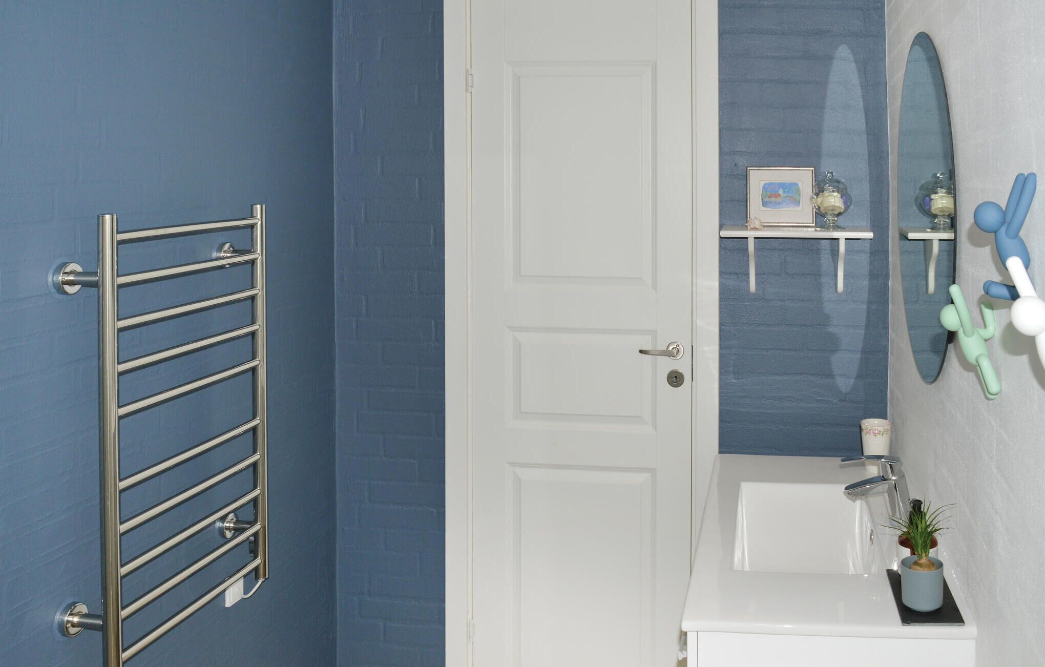 A bathroom with a towel rack, a door, a mirror, a sink, and a small shelf with decorative items.