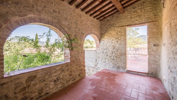 Une pièce rustique aux murs de pierre, dotée de fenêtres en arc de cercle et d’un sol carrelé, qui offre une vue sur la verdure et les collines.