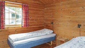A wooden cabin room with two single beds, a window with curtains, and a small wooden nightstand.