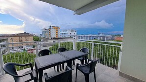 Een balkon met een tafel en stoelen, met uitzicht op stadsgebouwen en een verre kustlijn.