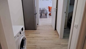 Een smalle gang met een wasmachine, een koelkast en een eethoek in de verte.