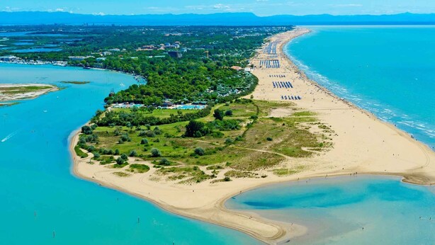 Luchtbeeld van een kustgebied met een strand, helderblauw water en een combinatie van natuurlijke en bebouwde gebieden.