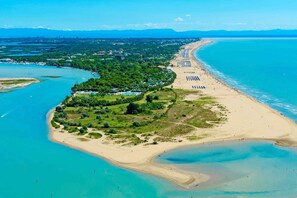 An aerial view of a coastal area with a beach, clear blue waters, and a mix of natural and developed land.