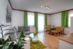 Un salon comprenant un canapé, une table basse en verre ornée de fleurs, une table  à manger avec des chaises, et un coin cuisine.