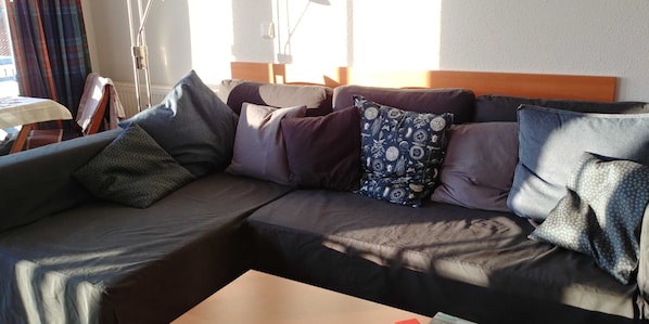 A dark-colored sofa with multiple pillows, a small table with a white tablecloth, and a window with curtains.