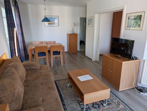 Una sala de estar amoblada con sofá, una mesa de centro, un televisor sobre un mueble de madera, una mesa de comedor con sillas y un tapete estampado.