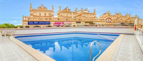 A swimming pool with a tiled deck, surrounded by a building with multiple balconies and a sign for 'The Bed Specialists'.