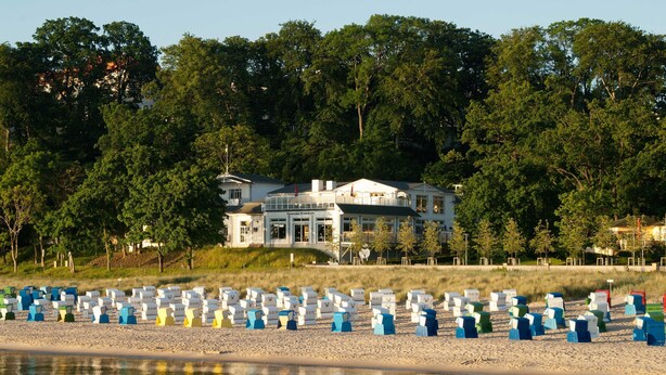 A beachfront property with a large house, a sandy beach with colorful beach chairs, and dense trees in the background. - Strandkorbhaus 9 150 m to the beach quiet location - Strandkorbhaus 9 150 m to the beach (Göhren)