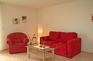 Uma sala de estar com uma poltrona vermelha, um sofá vermelho e uma mesa de centro de vidro. Há duas obras de arte penduradas na parede e um abajur de chão alto.