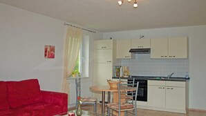 A compact living space with a red sofa, a glass coffee table, a dining table with chairs, a kitchen area with cabinets and a stove, and a window with curtains. - Strandkorbhaus 9 150 m to the beach quiet location - Strandkorbhaus 9 150 m to the beach (Göhren)