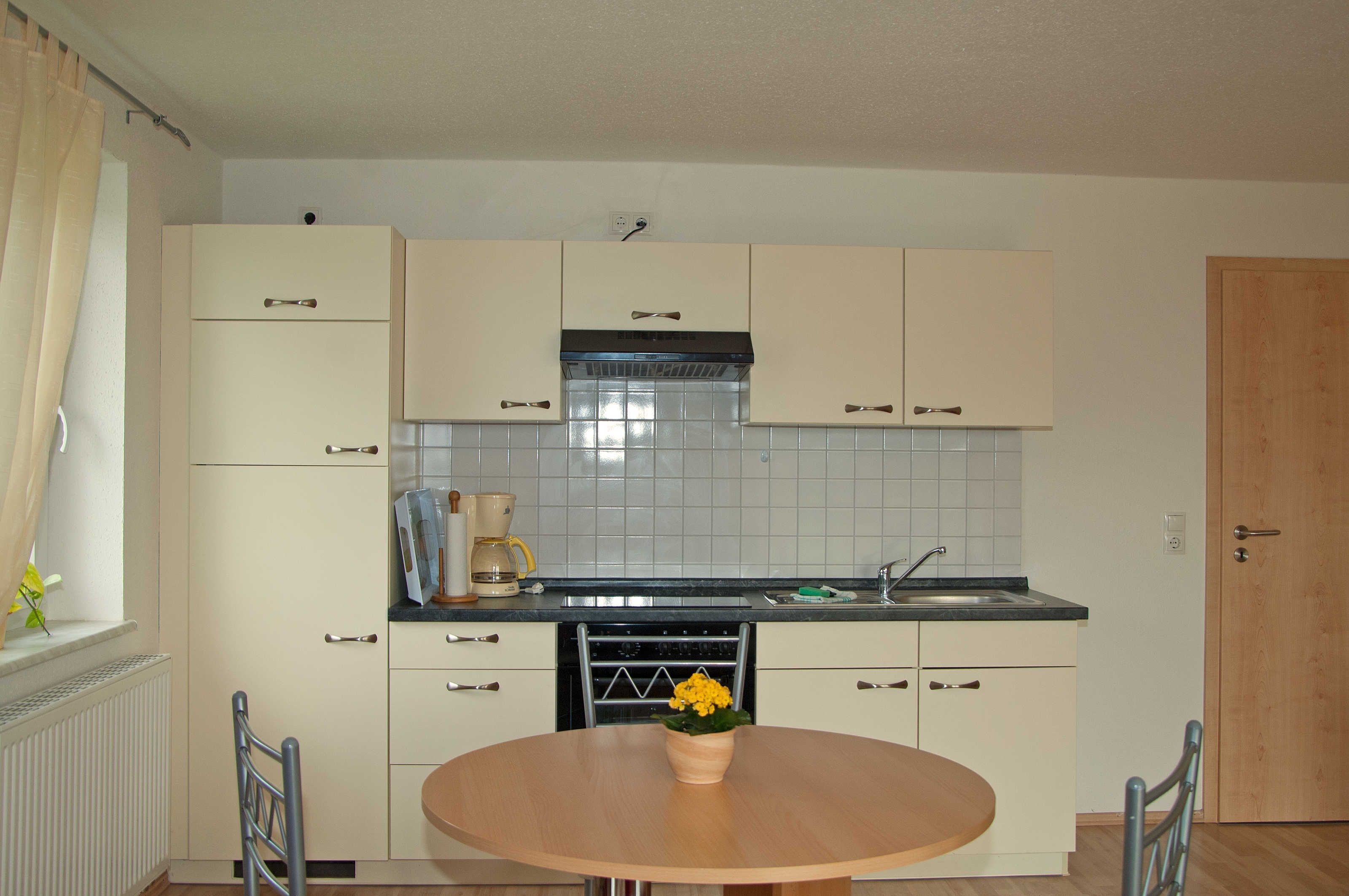A compact kitchen with white cabinets, a black range hood, and a tiled backsplash. There is a wooden dining table with a flower vase.