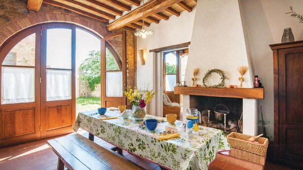 A rustic dining area with a wooden table set for two, a fireplace, and a view of the garden through large windows. - Cozy apartment in S. Quirico d'Orcia SI (S. Quirico d'Orcia SI)