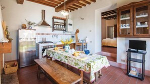 A compact kitchen with a stove, sink, and wooden ceiling beams. A dining table with a floral tablecloth is set in the center. - Cozy apartment in S. Quirico d'Orcia SI (S. Quirico d'Orcia SI)