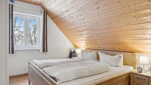 A bedroom with a sloped ceiling, a bed with white bedding, a bedside table with a lamp, and a window with curtains. - Ferienhaus 44 Altefähr (Altefähr/Rügen)