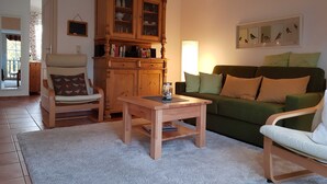 Una sala de estar acogedora con un mueble de madera, un sofá verde, una mesita de centro de madera y un sillón con cojín.