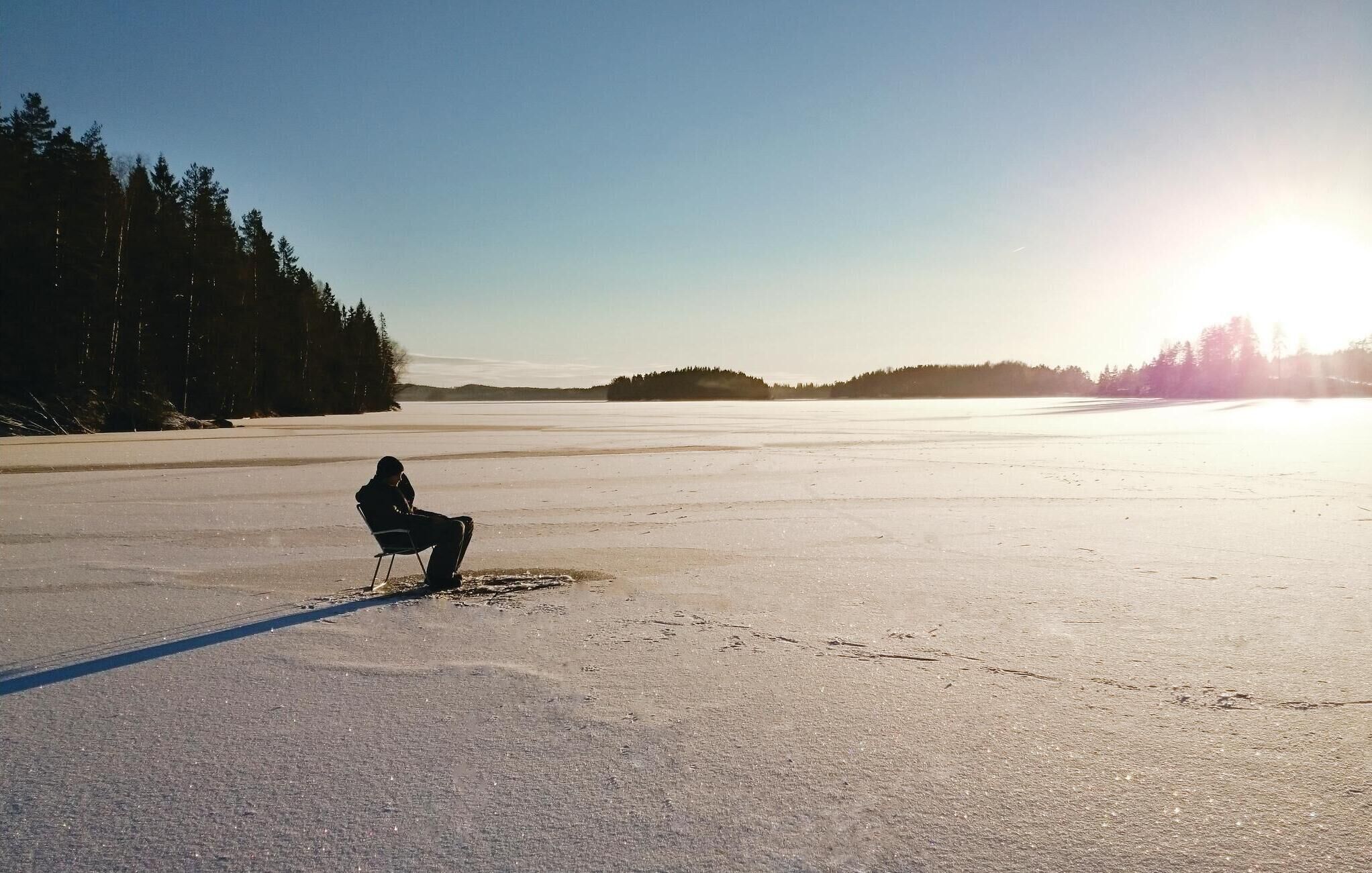 Eine Person sitzt auf einem Stuhl in einer weiten, offenen, verschneiten Landschaft mit blauem Himmel und Bäumen in der Ferne.