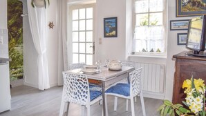 Un coin repas avec une table dressée pour quatre personnes, des chaises blanches avec des coussins bleus, un radiateur blanc placé sous la fenêtre et un téléviseur posé sur un meuble en bois.