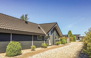 A row of cabins with dark siding and tiled roofs, arranged along a gravel path with small shrubs and a wooden fence in the background.