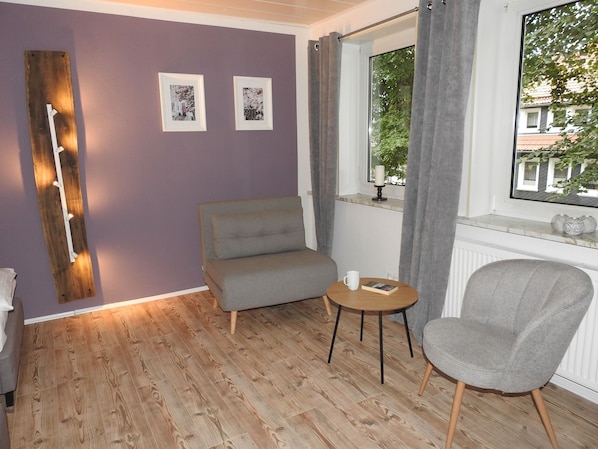 A room with a grey sofa, a round wooden table, a grey armchair, a window with curtains, and two framed pictures on the wall.