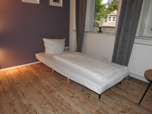 A single bed with a wooden floor, a window with curtains, and a radiator.