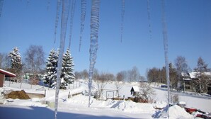 Un paisaje nevado con estalactitas colgando de un tejado, árboles cubiertos de nieve y un cielo azul despejado.