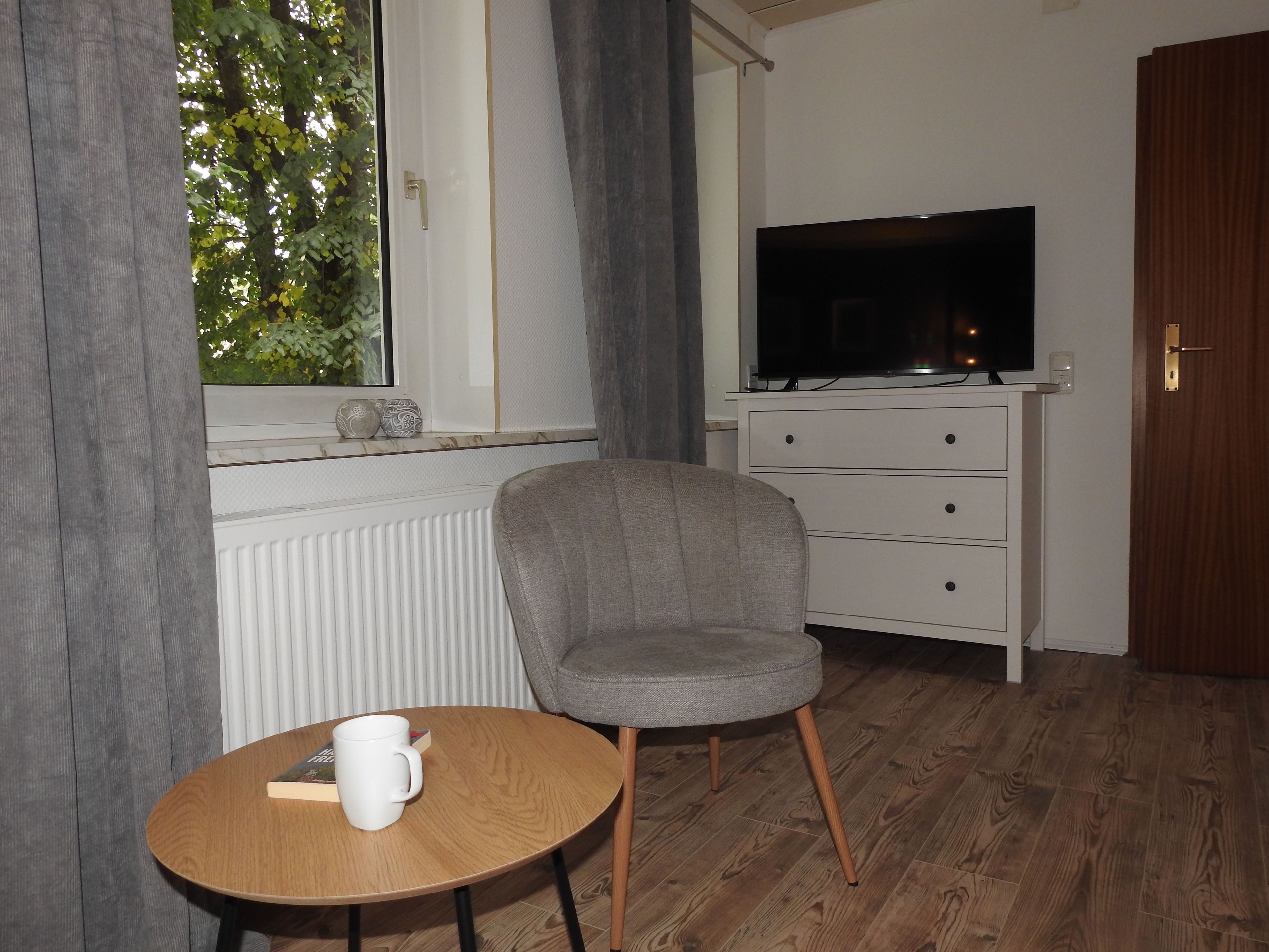Une pièce avec une télévision posée sur une commode, une chaise, une petite table avec un livre et une tasse, et une fenêtre avec des rideaux.