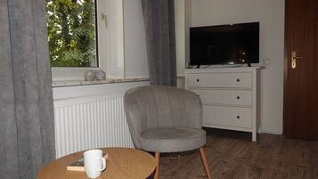 Une pièce avec une télévision posée sur une commode, une chaise, une petite table avec un livre et une tasse, et une fenêtre avec des rideaux.
