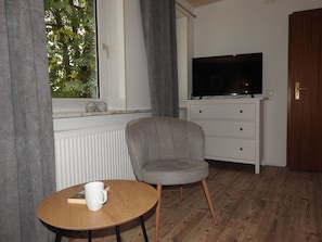 Une pièce avec une télévision posée sur une commode, une chaise, une petite table avec un livre et une tasse, et une fenêtre avec des rideaux.