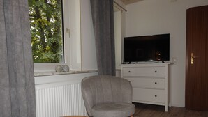 Una habitación con televisión sobre un aparador, una silla, una mesita con un libro y una taza, y una ventana con cortinas.