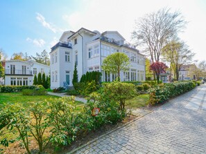 A multi-story white building with a balcony, surrounded by well-maintained gardens and a cobblestone pathway.