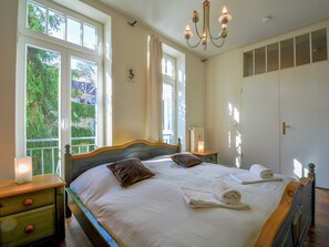 Ein Schlafzimmer mit einem großen Bett, einer Chandelüster, einem Fenster mit Blick ins Grüne und einer Tür.