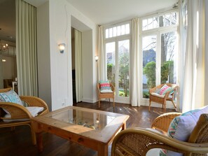 A room with wicker furniture, a glass-top table, and large windows with curtains.