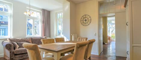A dining area with a wooden table and chairs, a clock on the wall, and a view of the outside through large windows.