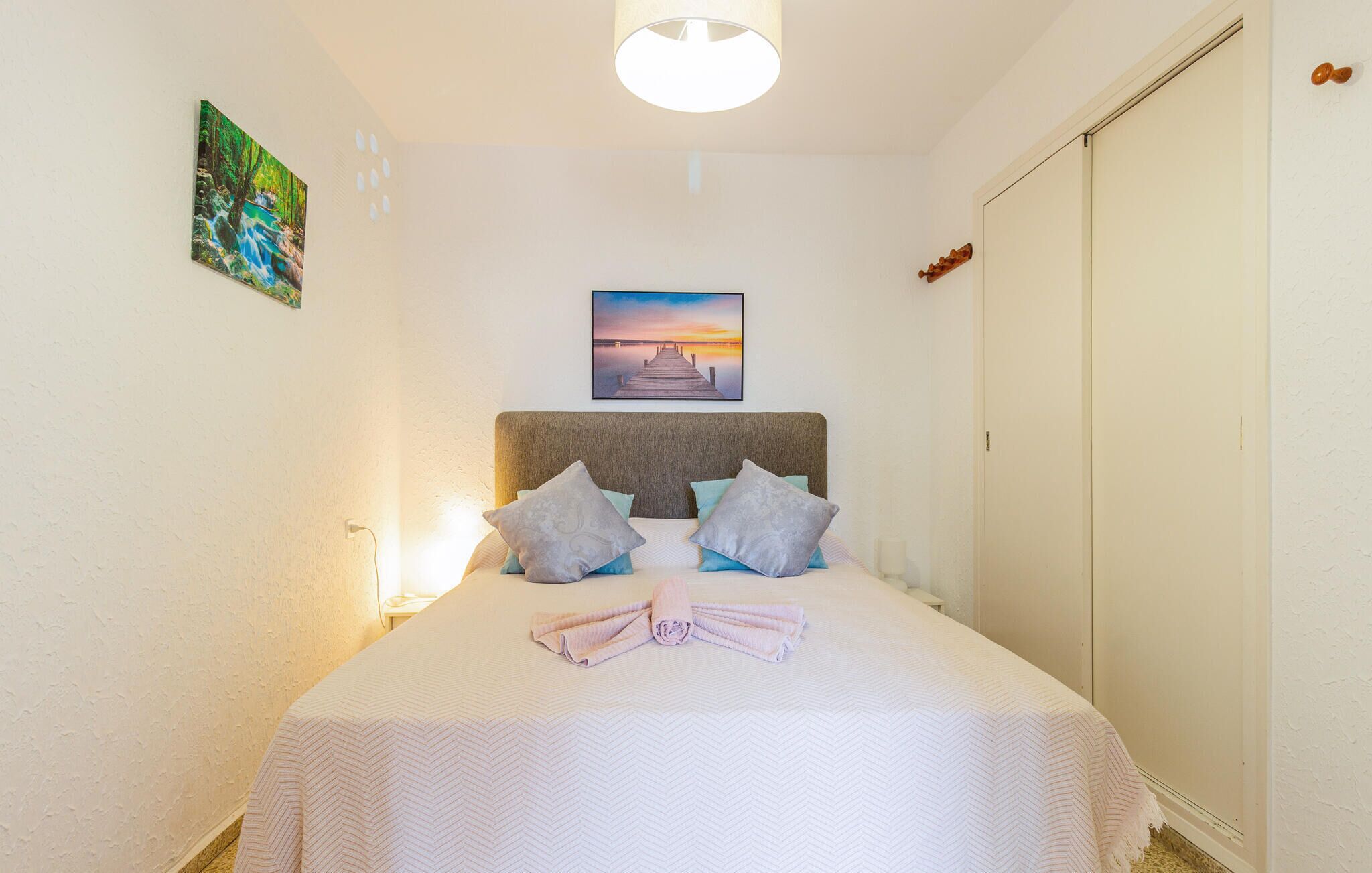 A neatly made bed with a white bedspread and two pillows, a painting of a sunset on the wall, and a wooden door to the right.
