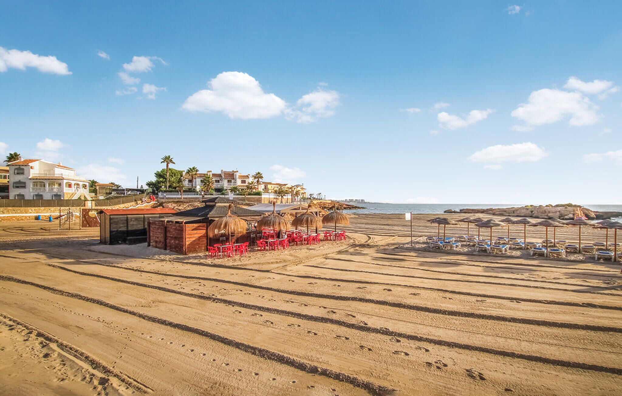 Une plage avec des chaises et des parasols rouges, un petit bâtiment et des maisons en arrière-plan.