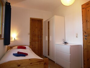Ein kleiner, gemütlicher Raum mit einem hölzernen Bett, einem weißen Kleiderschrank und einem kleinen Schreibtisch mit Lampe.