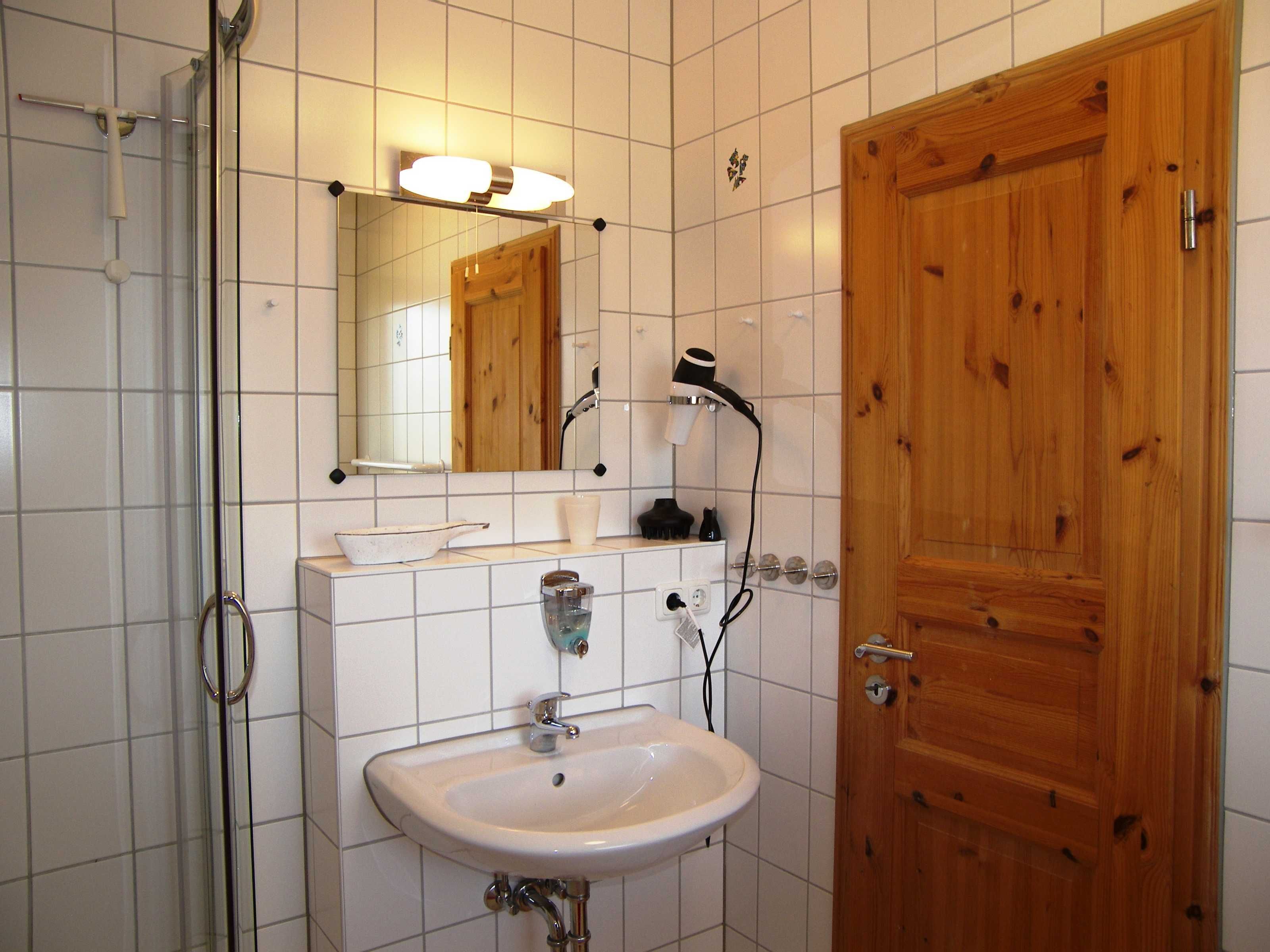 Ein Badezimmer mit weißem Waschbecken, einem Spiegel, einer Holztür und einer Dusche.