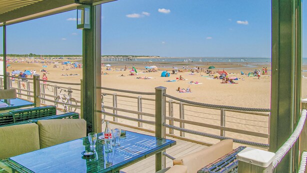 A beach view from a covered area with tables and chairs, a railing, and a view of the beach with people and umbrellas. - 3 bedroom lovely home in La Tranche sur Mer (La Tranche sur Mer)