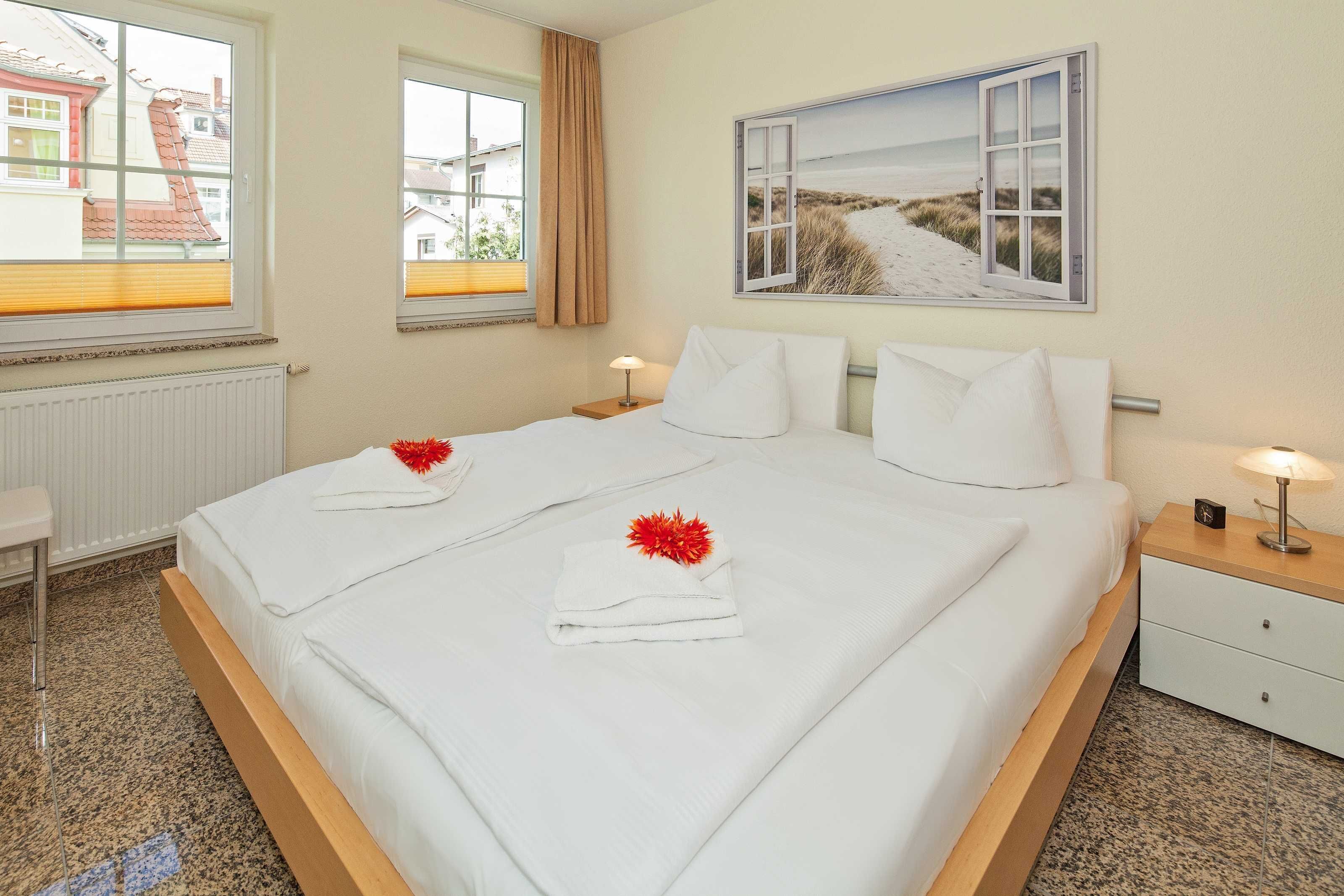 A neatly made double bed with white linens and red flowers, a bedside table with a lamp, and a window with a view of buildings.
